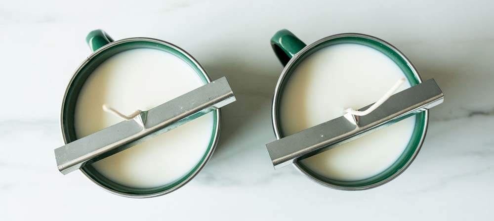 Overhead of two enamel mug candles cooing with the wick bars holding the wicks in place.