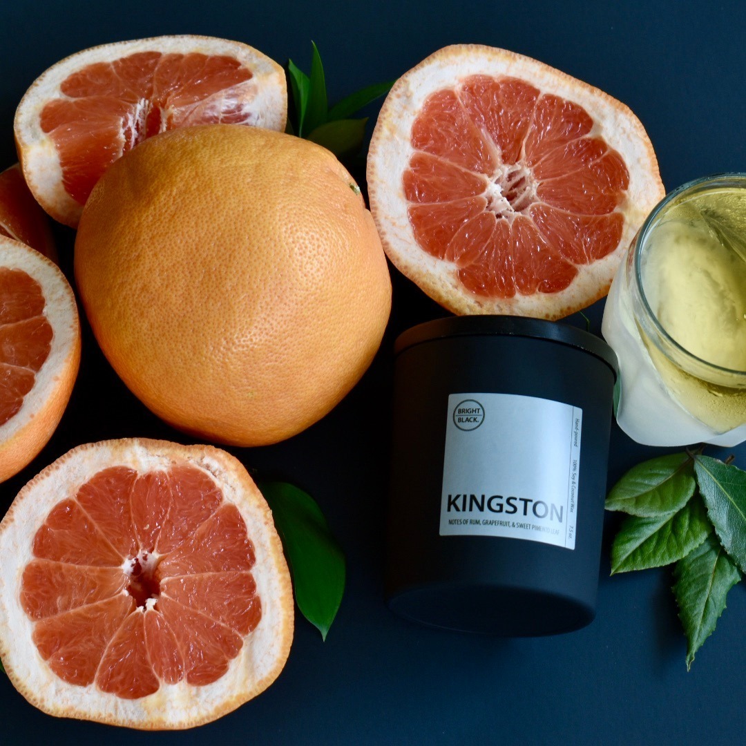 A candle with a black container and black lid labeled Kingston lies beside slices of grapefruit and green leaves.