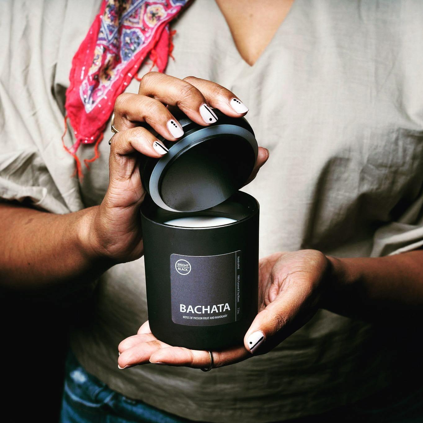 A woman holding a candle with a black container and black lid and label that says Kingston. She holds the lid open to reveal off-white wax.