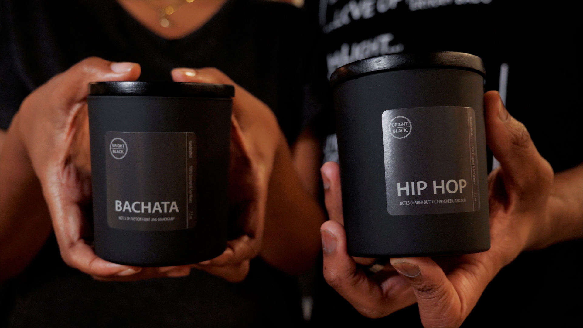 Hands holding candles with black containers and black lids. The candles are labeled Bachata and Hip Hop.