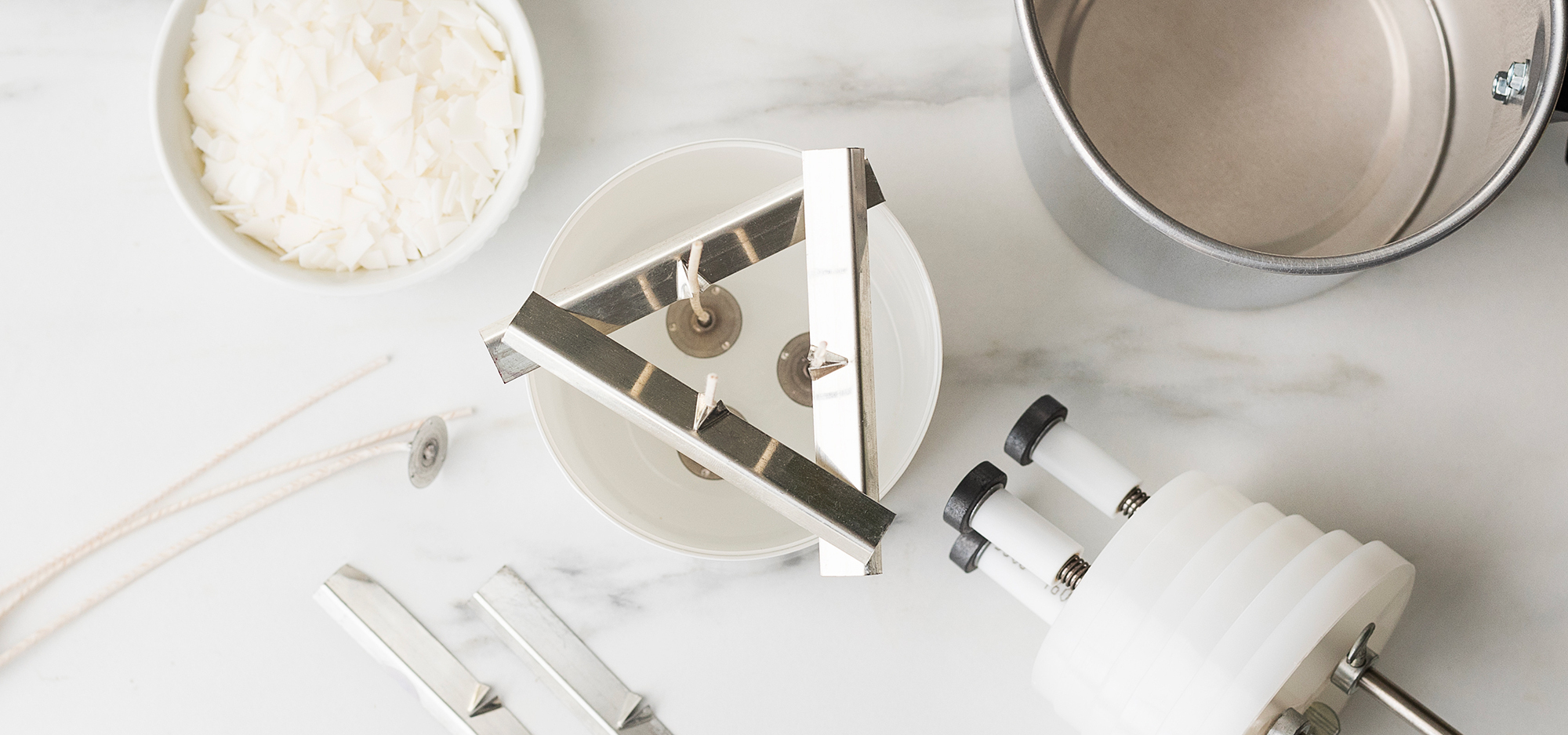 Candle making supplies on a marble surface, including a white candle container with three wicks held in place by silver wick bars, soy wax flakes in a white bowl, a silver pouring pitcher, and a wick-setting tool.