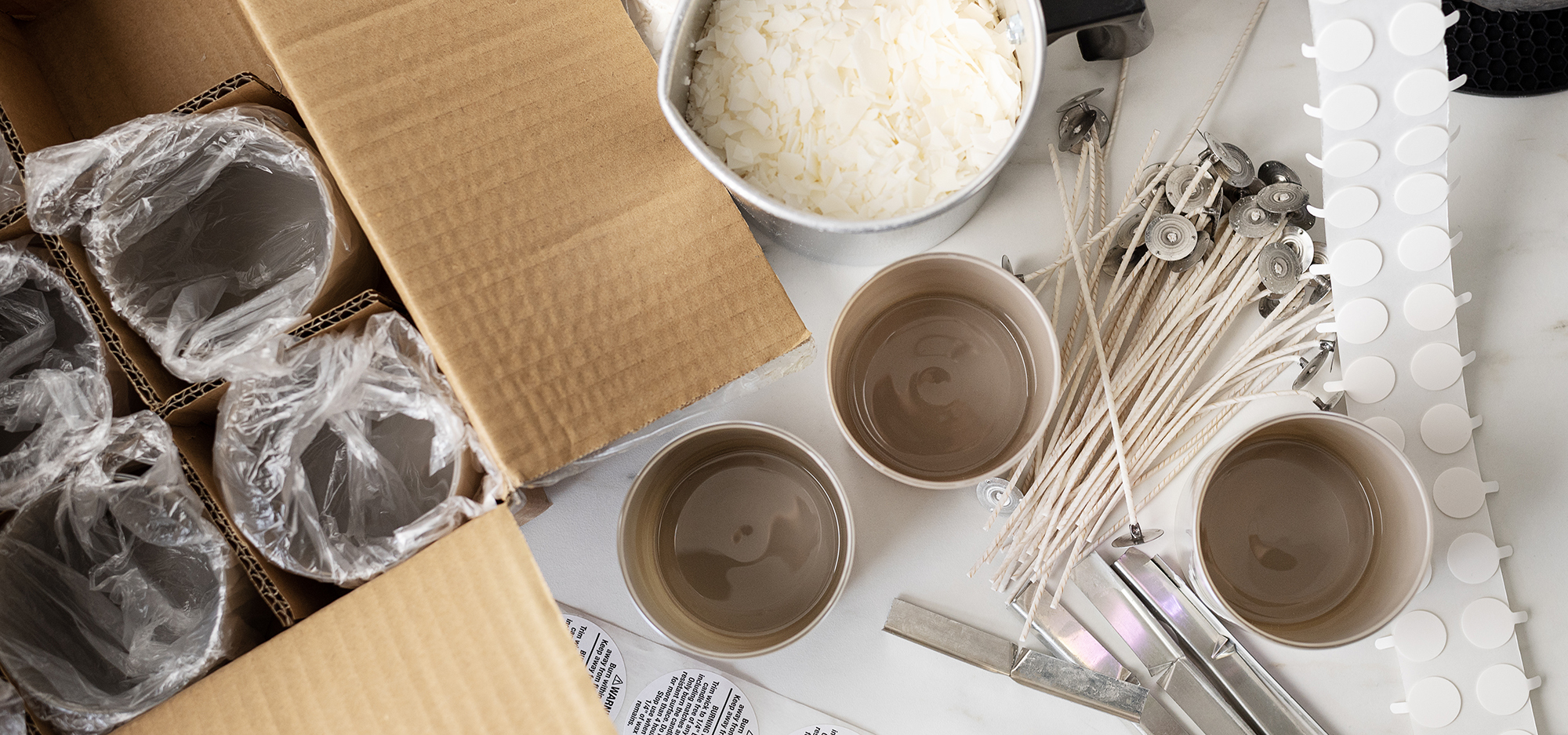 An array of candle making supplies: a cardboard box filled with empty candle jars, a metal pouring pitcher of soy wax flakes, candle wicks, candle bars, warning labels and a roll of wick stickers