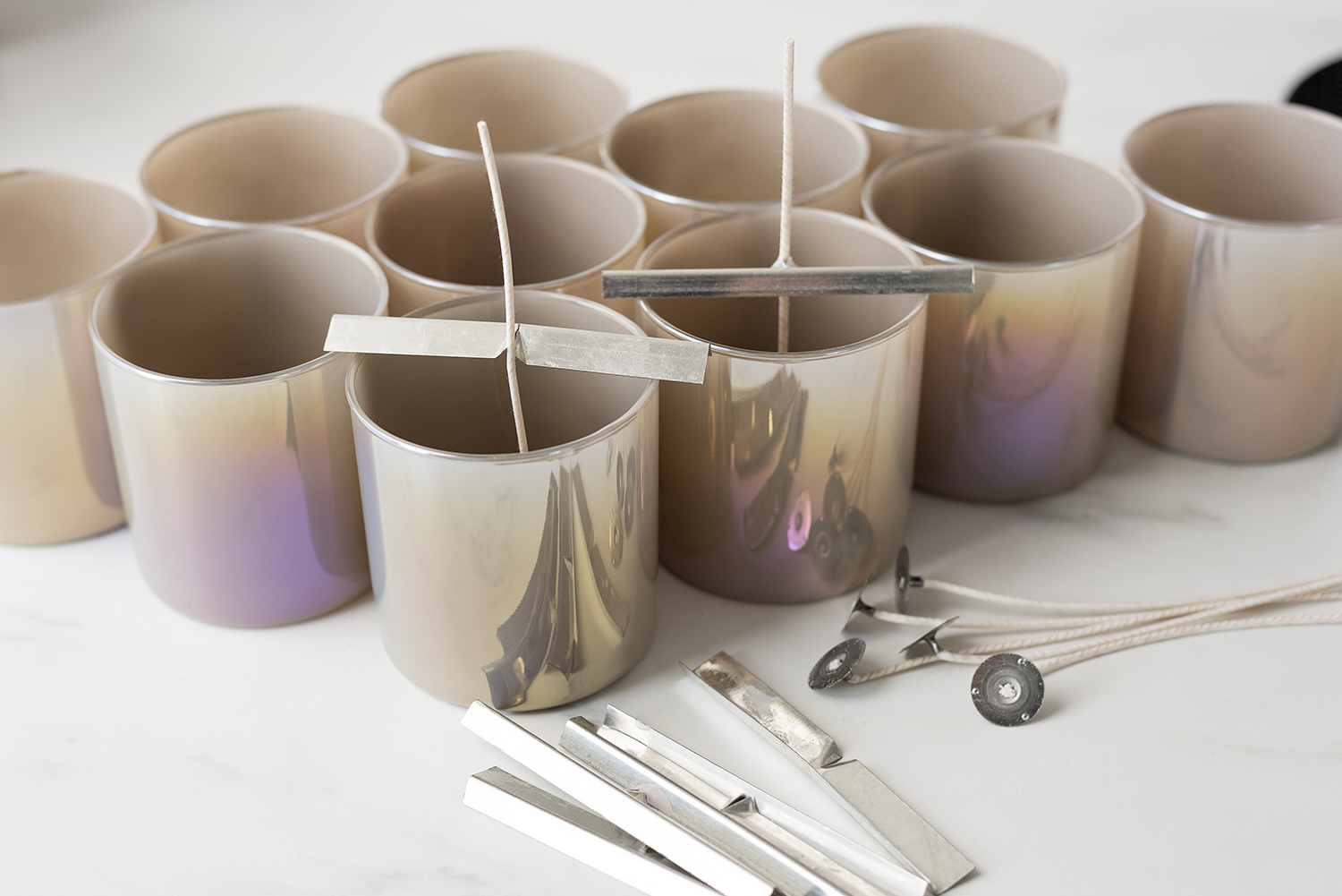 Three rows of taupe iridescent candle jars, candle wicks and wicks bars on a marble surface