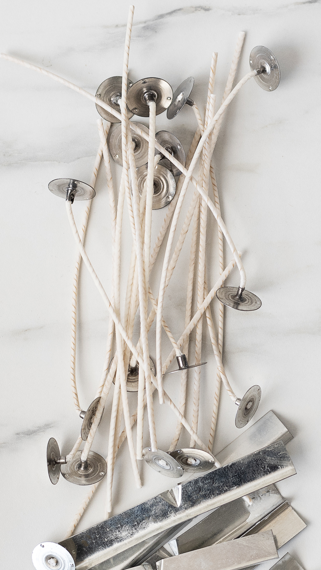 Groupings of pretabbled cotton candle wicks and silver-colored metal wick bars on a marble background