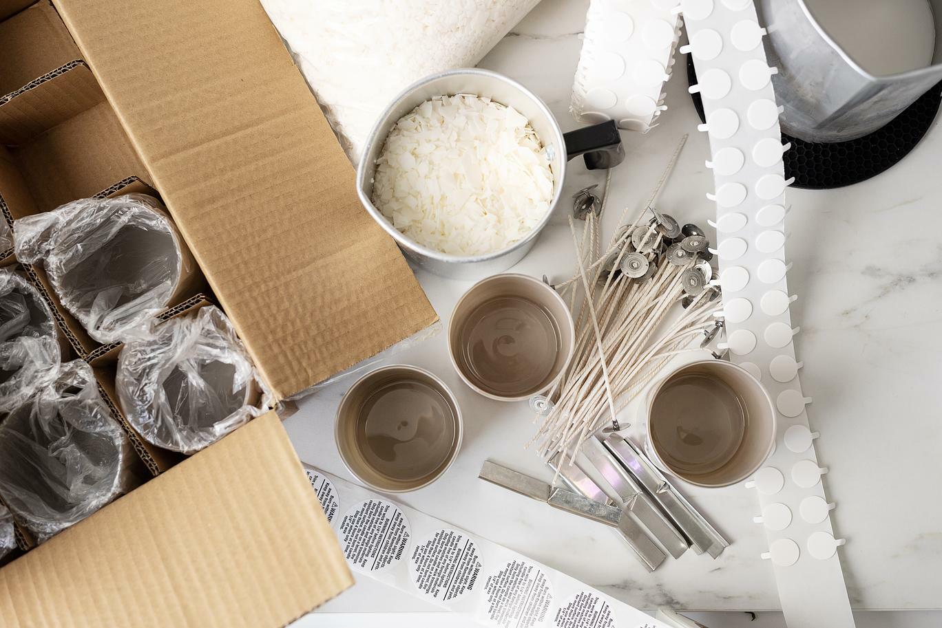 An array of candle making supplies: a cardboard box filled with empty candle jars, a metal pouring pitcher of soy wax flakes, candle wicks, candle bars, warning labels and a roll of wick stickers