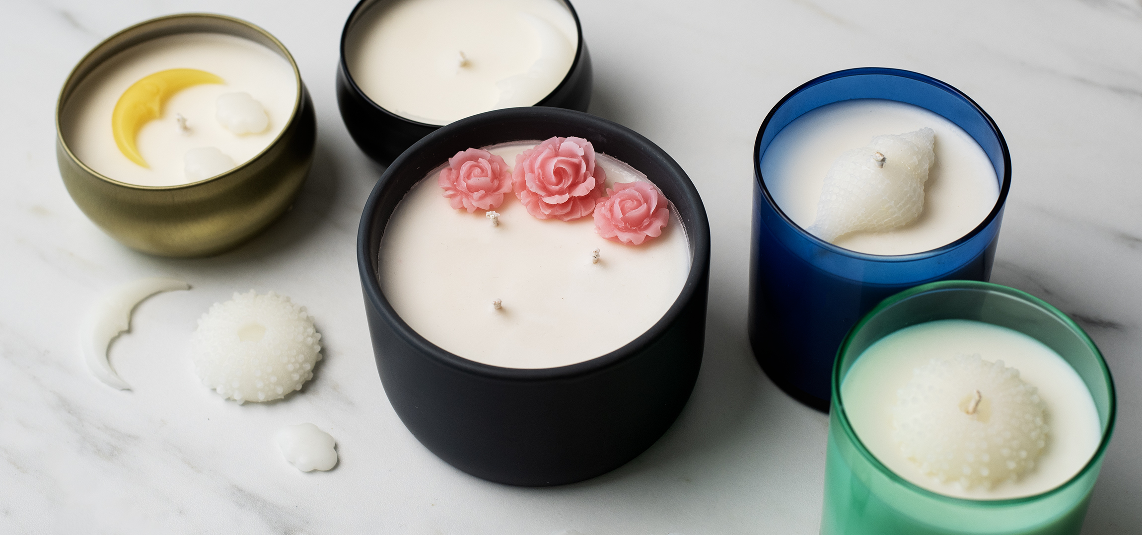 Five candles with decorative, 3-D wax embeds on their surface. The wax embeds are shaped like crescent moons with faces, clouds, roses, and seashells. The candle containers are metallic gold, black, blue, and teal. 