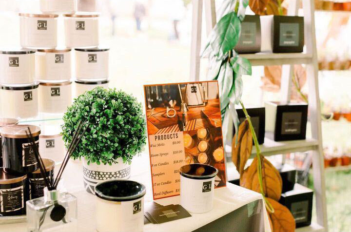 In this brightly lit image, candles in white and black straight sided glass tumblers are displayed at a craft show. A color-printed 8-1/2"x11" vertical sign displays product prices. A white shelf of candles in black boxes is out of focus in the background.