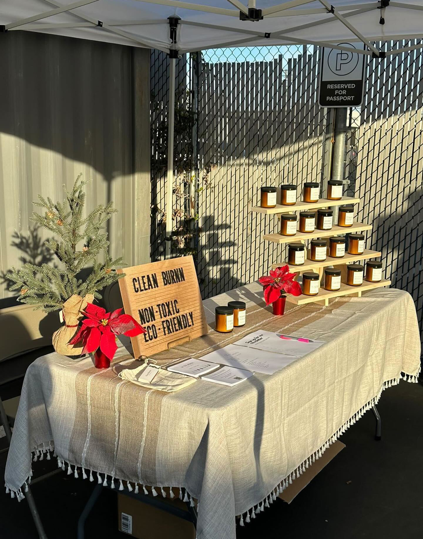 Under a white canopy tent sits a table covered in a linen table cloth. Displayed on the tabletop is a four-level natural wood riser with 16 candles in amber glass jars. To the left of the display are poinsettias, a natural-colored wood letterboard, and printer collateral materials on the table.