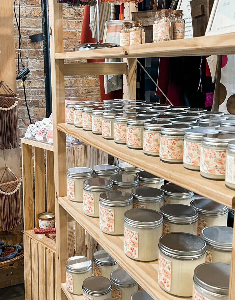 Four shelves of candles in small and medium straight sided clear glass jars with silver lids are displayed at an indoor market on a shelf made of natural wood