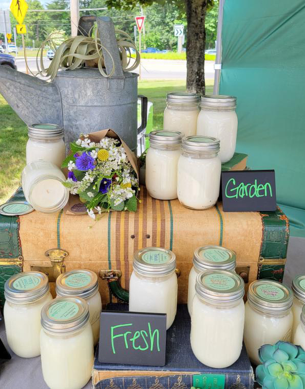 More than a dozen soy candles made in clear glass 16 oz. mason jars with silver lids are displayed at an outdoor market. A tan vintage suitcase serves as the display piece with candles in front of and on top of the suitcase. Small black chalkboard signs feature the words Garden and Fresh in green chalk pen. A galvanized metal vintage-style watering can and a bouquet of flowers wrapped in brown paper accent the display.