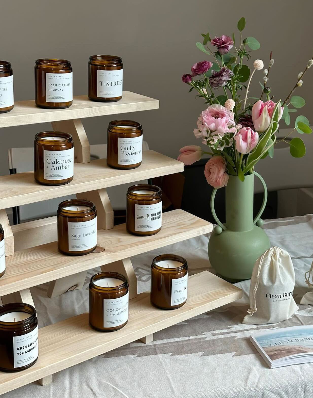 In this cropped image, a four-shelf rise made of natural wood displays soy wax candles made in amber glass jars, shown without lids. To the right of the display is a shapely matte sage green vase with delicate pink and purplee flowers. In front of the vase is a muslin bag, likely containing a candle, stamped with the Clean Burnn brand name. A stack of cards with the Clean Burnn name are in the foreground. The tabletop is covered in a light tan tablecloth with a Native American motif.
