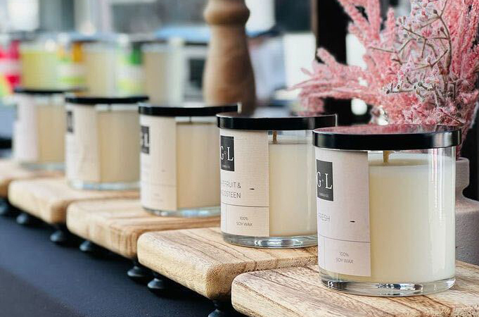 Soy wax candles in clear glass straight sided tumbler jars with black lids sit in a row on table draped in a black tablecloth. Each candle sits on its own natural wood-colored square riser. A sprig of pink foliage is out of focus in the background.