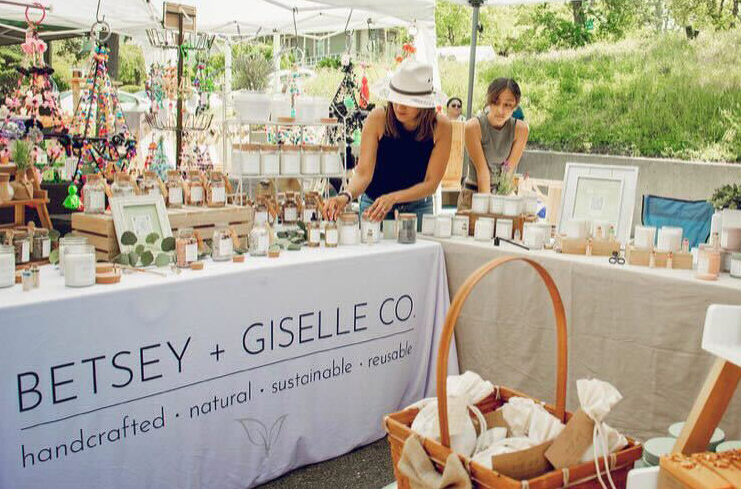 A light-skinned woman wearing a navy tank top and white hat stands behind two tables displaying handmade candles at an outdoor market. She is looking down while she arranges the candles. To her left is a young girl looking on. Their tables, draped in tablecloths of white and linen-colored fabric, are covered in candle; some are elevated on wood crates, small wooden cubes, and white tabletop metal shelves.