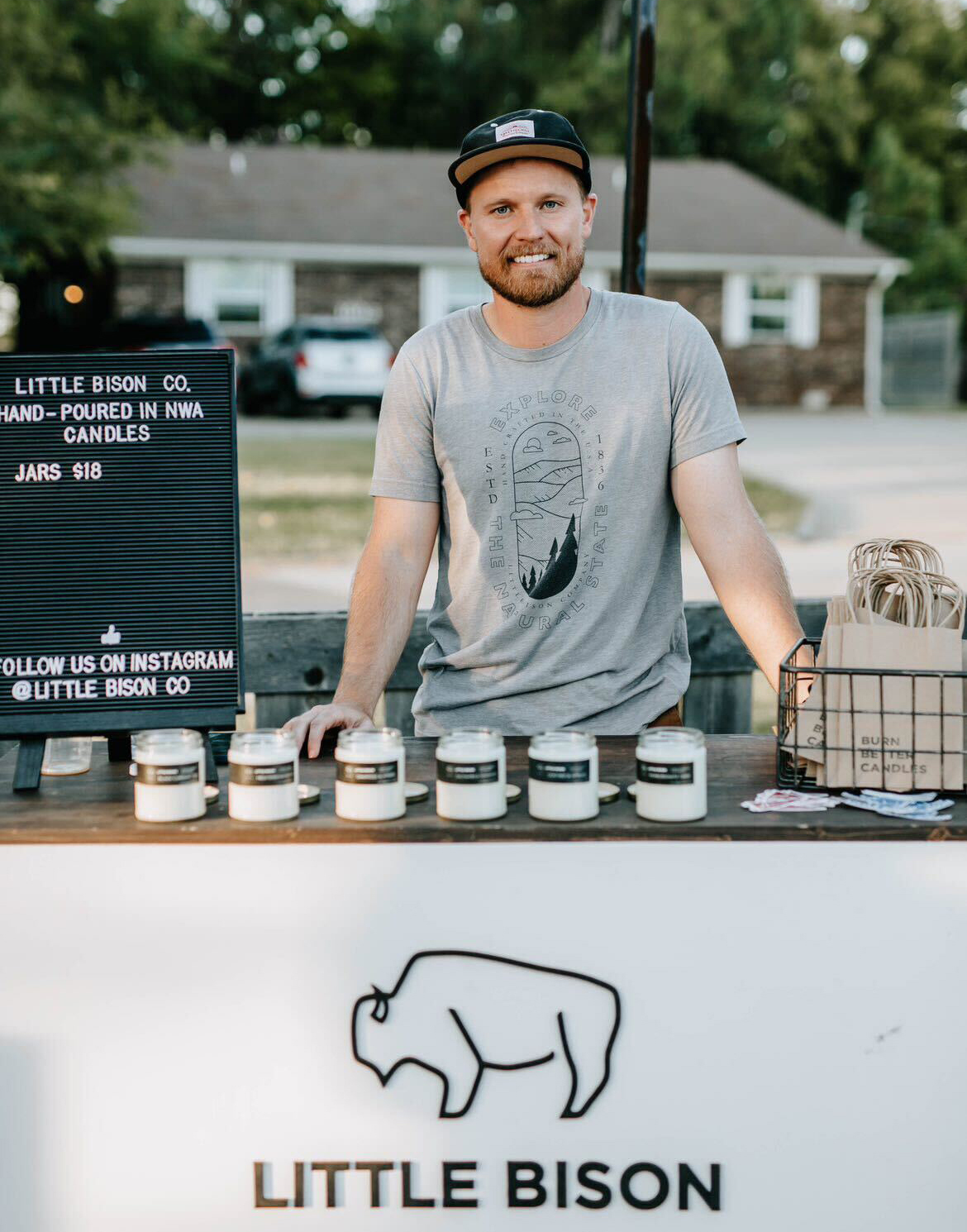 A smiling man with light skin looks directly at the camera while smiling. He stands behind a white cart that features a bison symbol and the words Little Bison. The cart is topped in dark-stained wood. A black letterboard is to the man's left. In front of him is a row of candles poured in clear glass straight sided jars with black labels. To his right is a black wire basket with kraft paper bags.