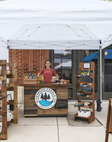 A woman wearing a read shirt stands proudly with her hands on hips behind a wood-clad counter under a white canopy tent. She looks directly into the camera and smiles.  Flanking her are rustic wooden shelves that display candles.