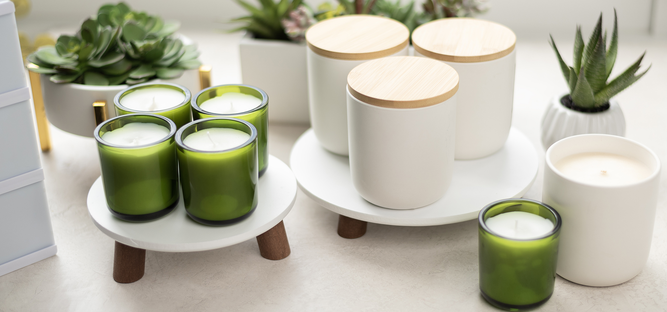Soy wax candles made in translucent green glass Sonoma jars and white ceramic Nordic tumblers with wood lids are arranged on white risers on a marble surface, and are surrounded by succulents in white pots
