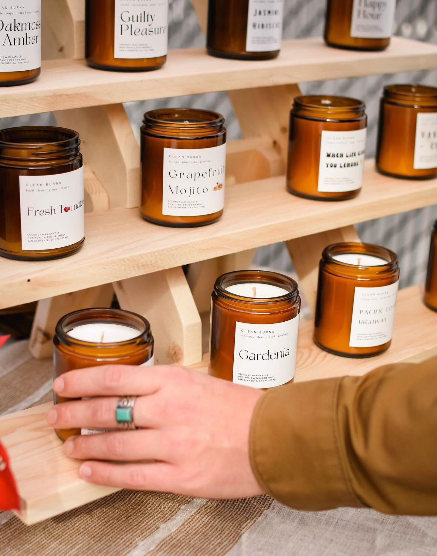 A light-skinned hand reaches for a single candle in a display of soy wax candles poured in amber glass jars with white labels. There are three rows of candles with their lids off on a natural wood riser.