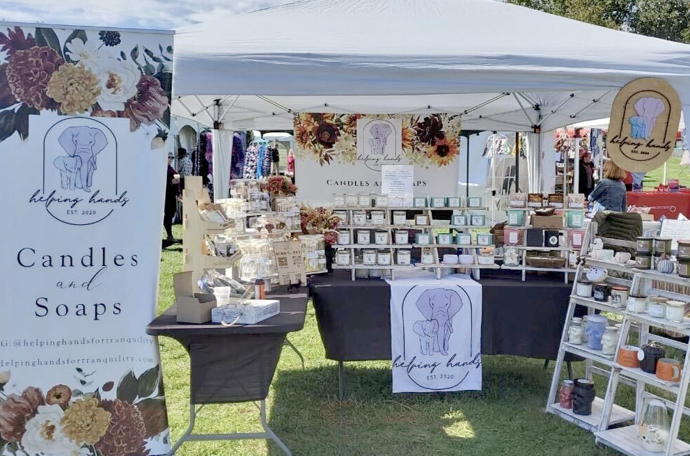 In this outdoor image, a vertical vinyl sign advertising candles and soaps is positioned in the left foreground; behind the sign is a white canopy tent with tables of candles and soaps displayed on risers; to the right are white shelves displaying more products for sale