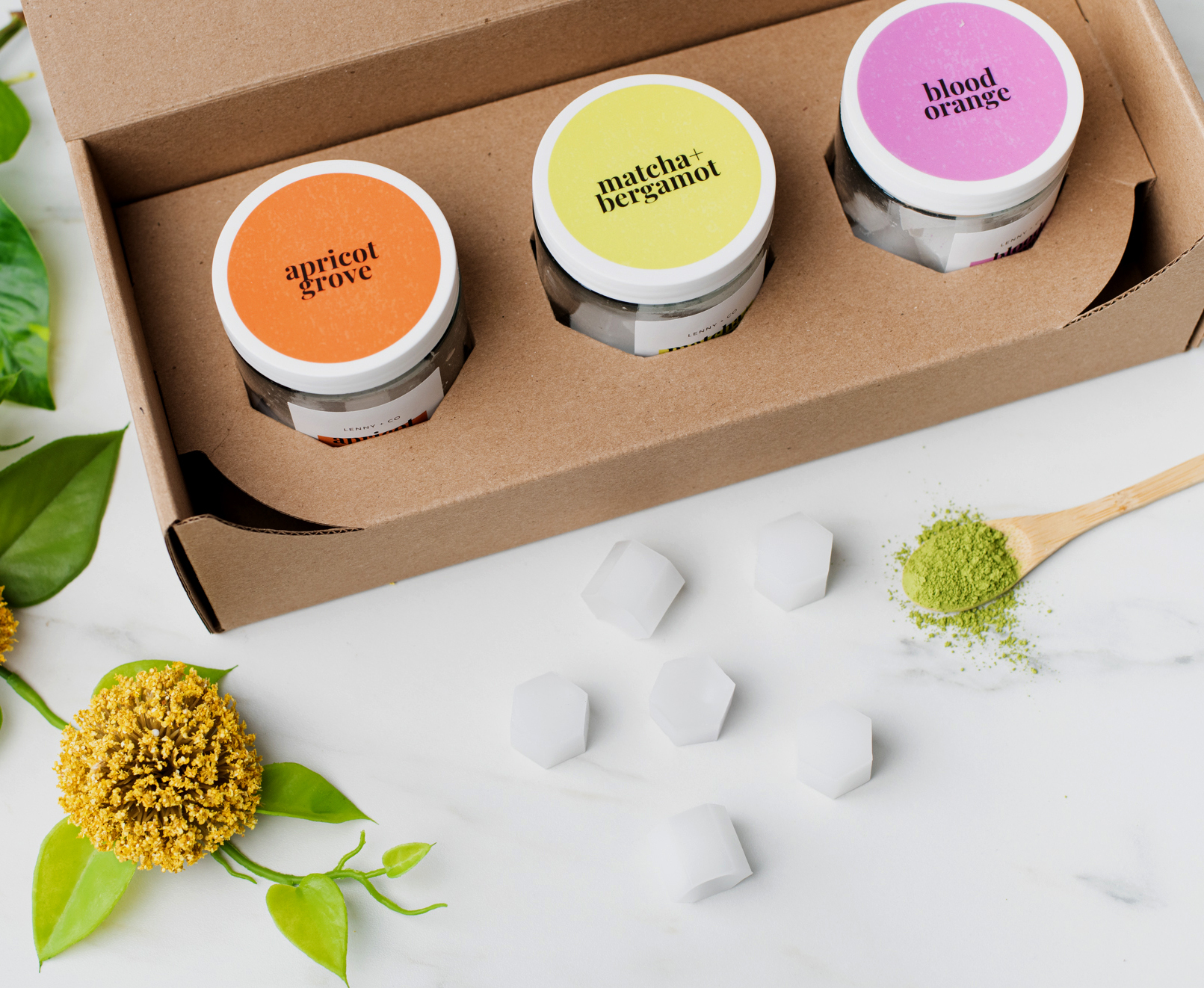 Orange, green, and purple labeled jars in a cardboard box on a white marble surface. Flowers, white wax melt squares, and matcha powder is beside the box. 