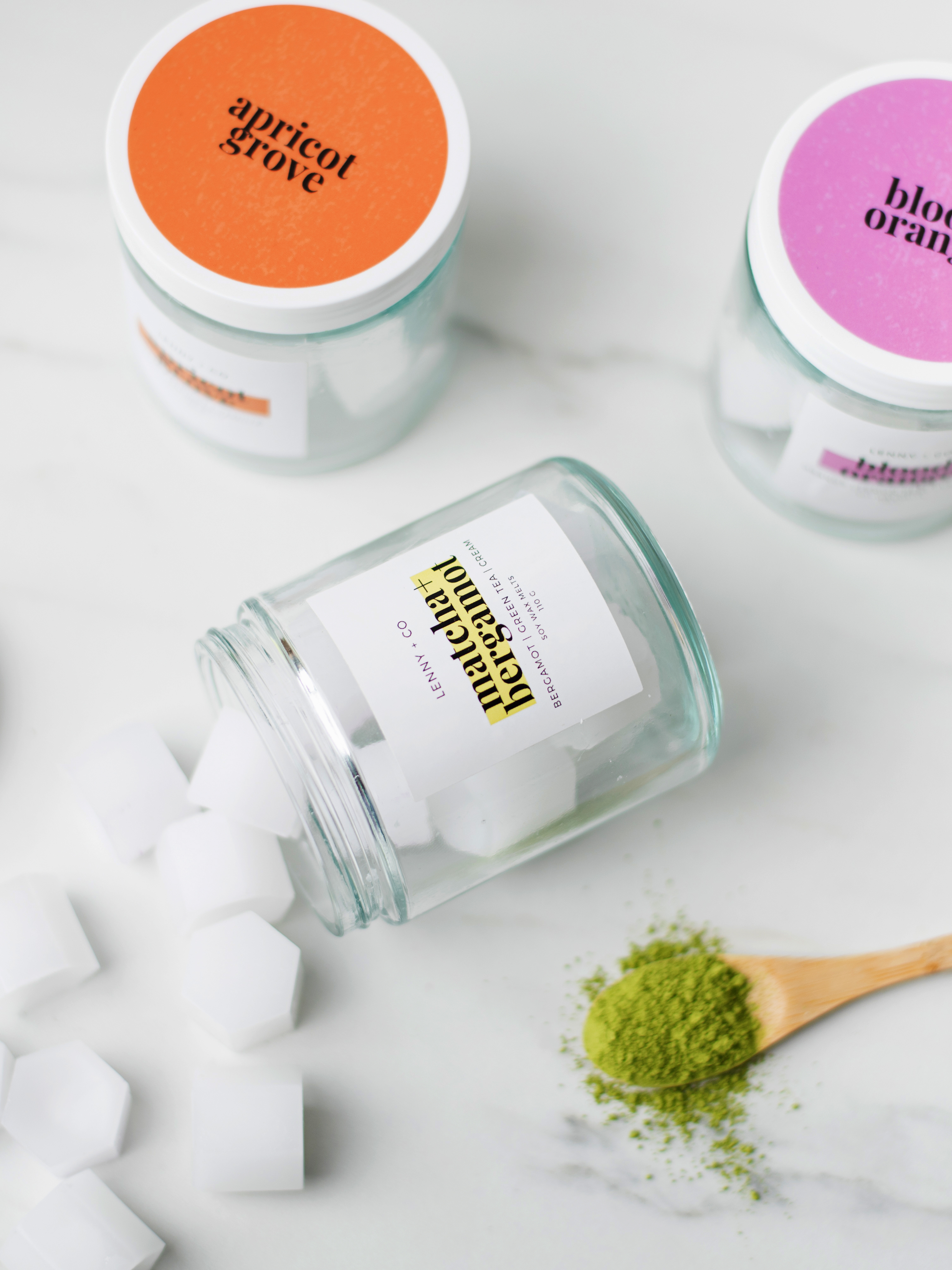 A jar with an orange label and a jar with a purple label sit upright beside a jar that's on its side. White square wax melts spill out of the jar on its side. A wooden spoon with matcha powder is towards the bottom of the photo.