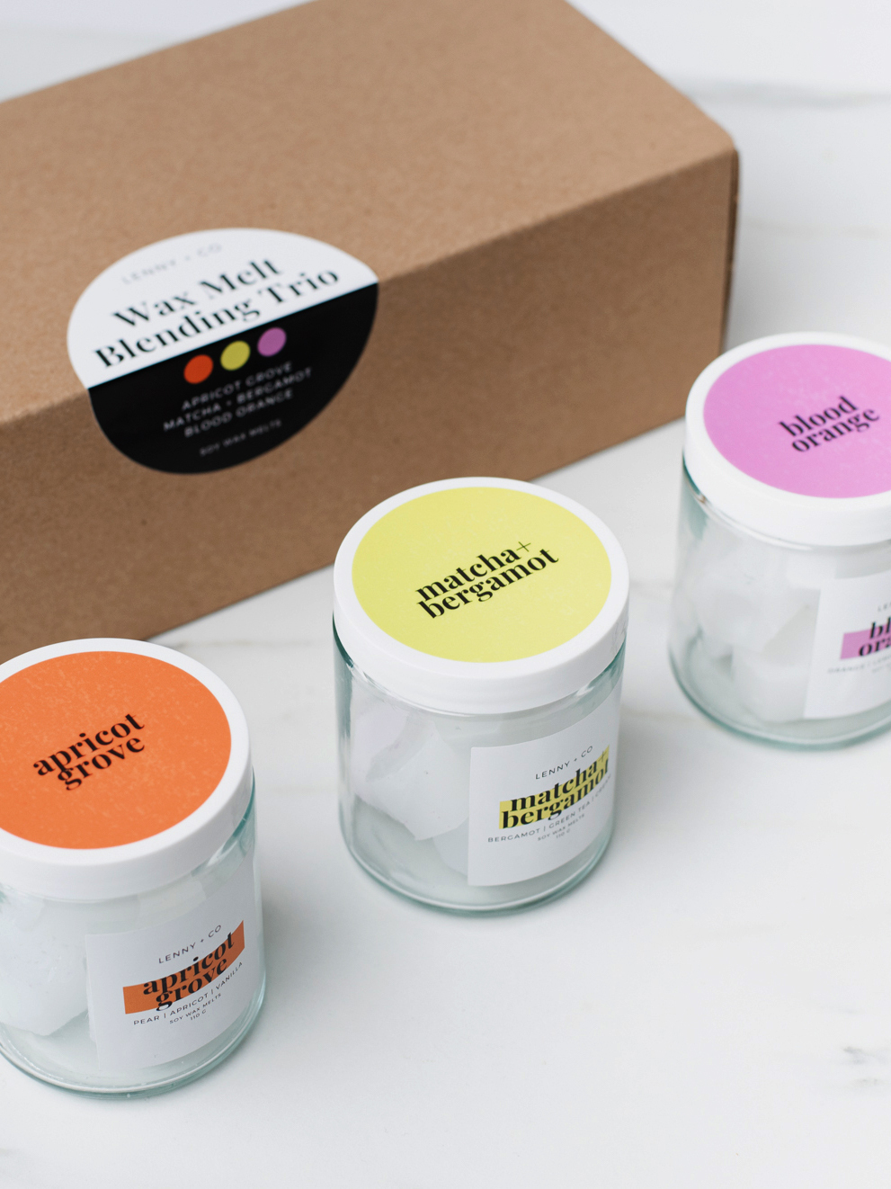 Three jars with orange, yellow, and purple labels are placed beside a closed cardboard box. There is a label that says "wax melt blending trio" on the closed box. 