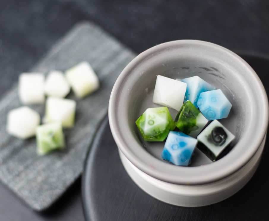 White, green, blue, and black wax melts shaped like dice in a circular, white ceramic wax melt warmer.
