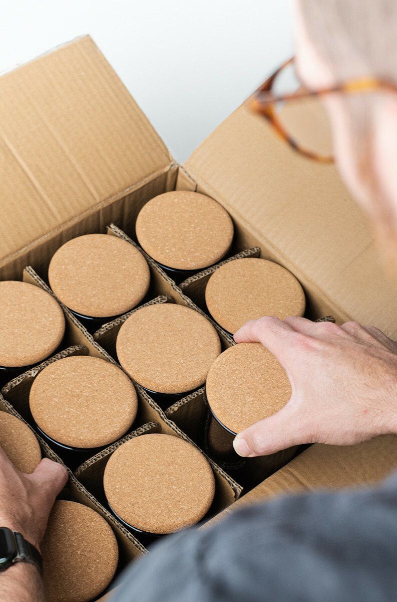 A light-skinned man with glasses reaches into a divided cardboard box filled with candles. The candles are topped with cork lids.