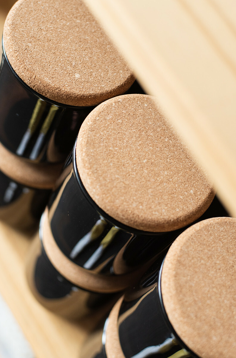 An angled overhead view of stacked rows of candles in black jars with cord lids on a natural wood shelf