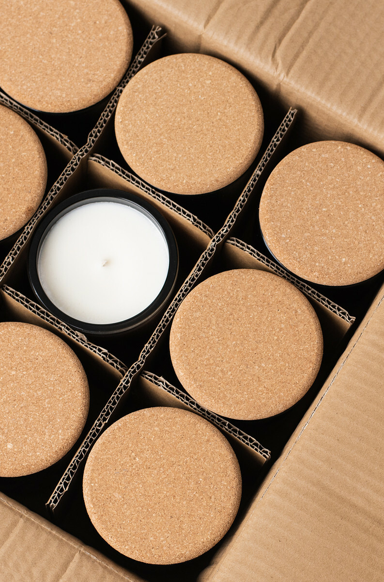 Overhead view of a divided cardboard box containing candles poured in black glass jars and topped with cork lids. A candle jar in the middle of the box is without a lid and displays surface and wick of the white candle.