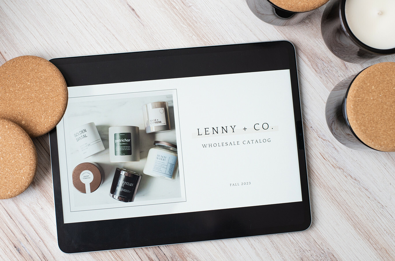 An iPad displaying a candle wholesale catalog cover sits on whitewashed wood surface with candles in black jars with cork lids surrounding it