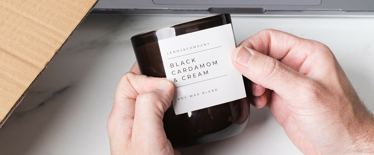 A pair of hands affixes a white label with black print to a black glass candle jar