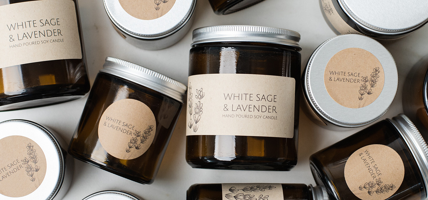 A flat lay of candles in amber glass jars with silver metal lids and kraft paper labels; some jars are on their sides while others are upright