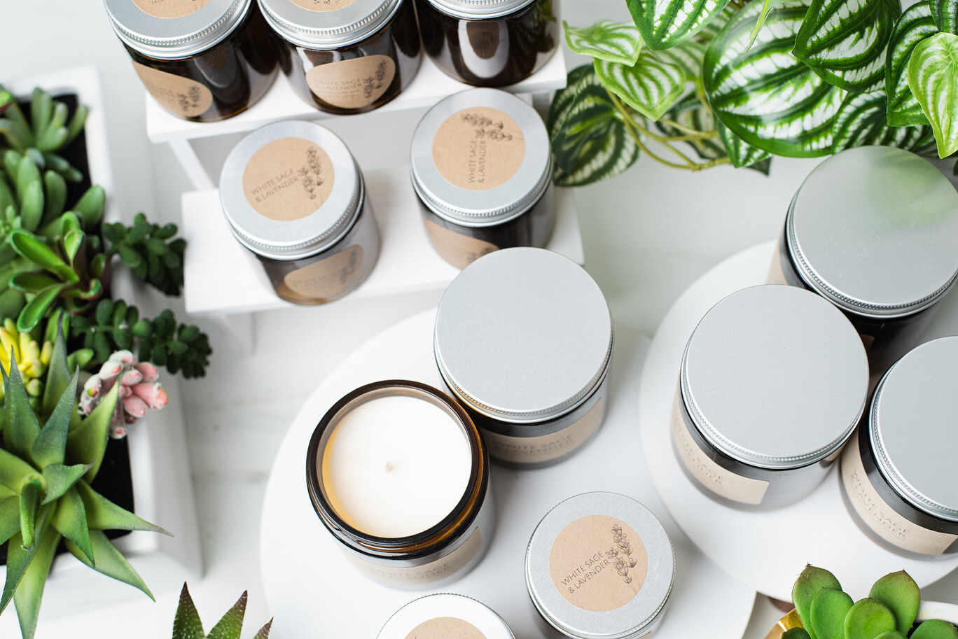 Overhead view of amber glass candle jars with silver metal lids on white risers surrounded by greenery