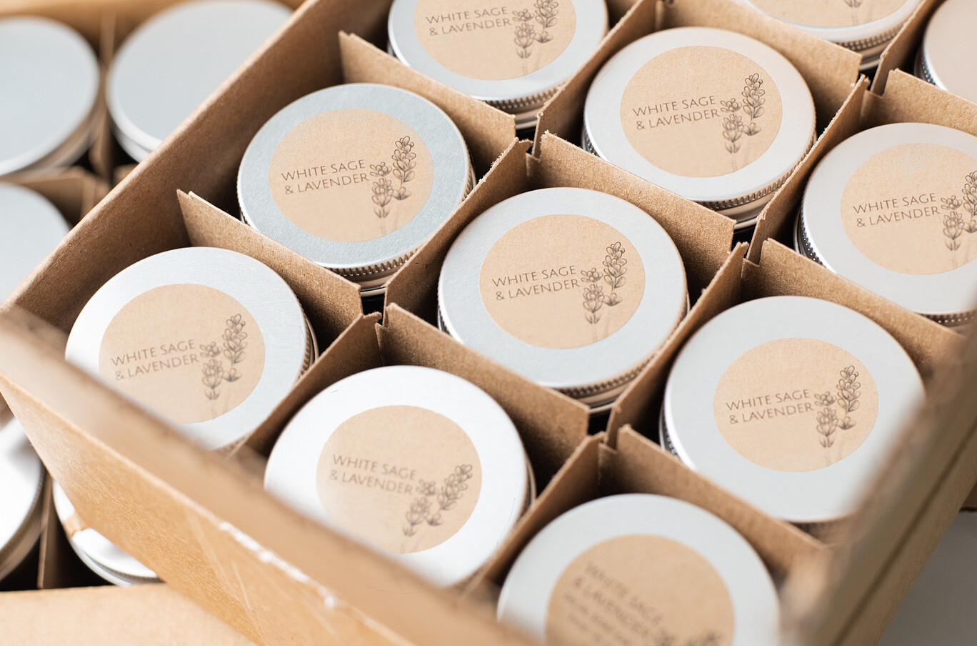 An angled overhead view of white label and private label candles packed in cardboard boxes with dividers; the candles are in amber glass jars with silver metal lids and kraft paper labels