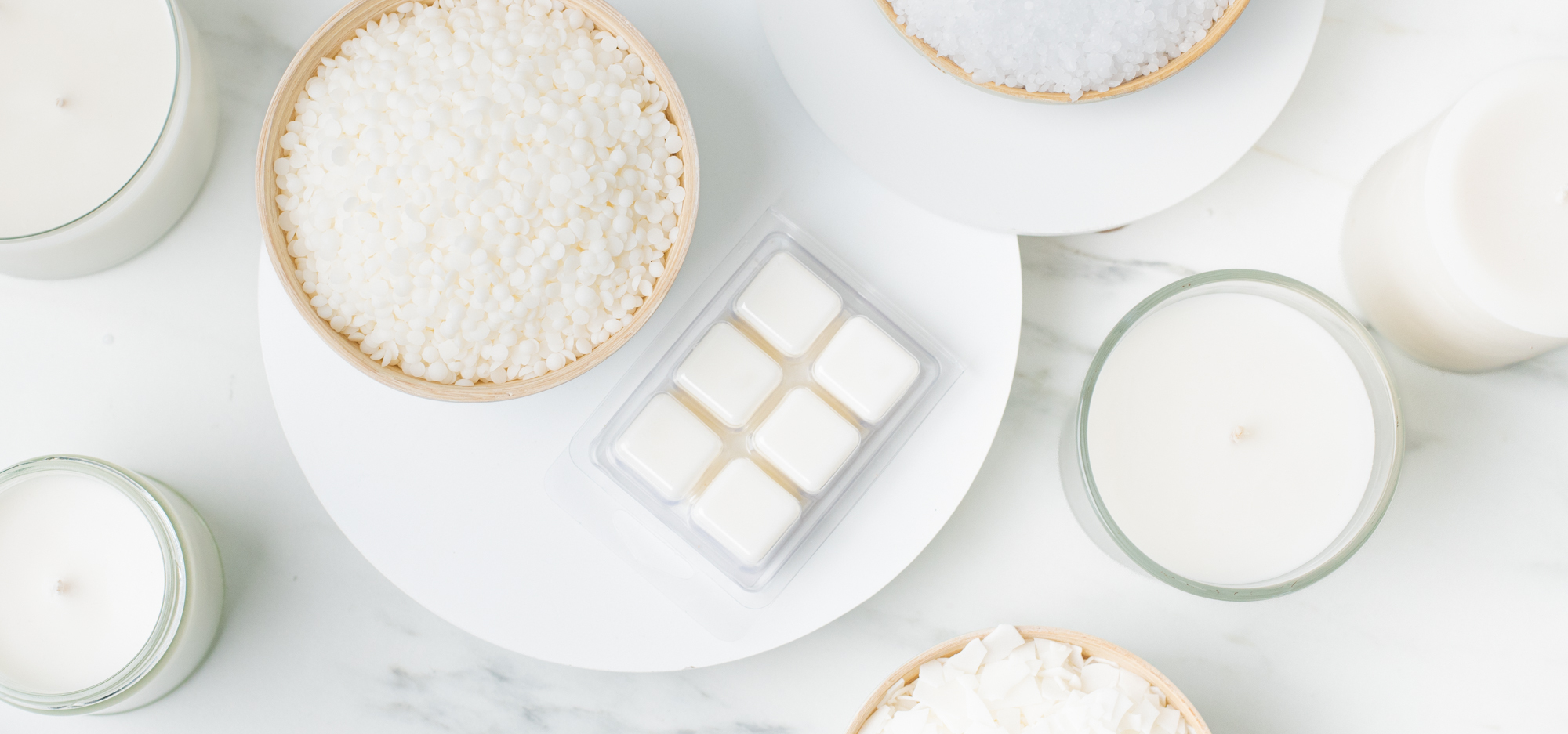 A white marble surface with bowls of unmelted wax pastilles, a clamshell wax melt, and two candles.