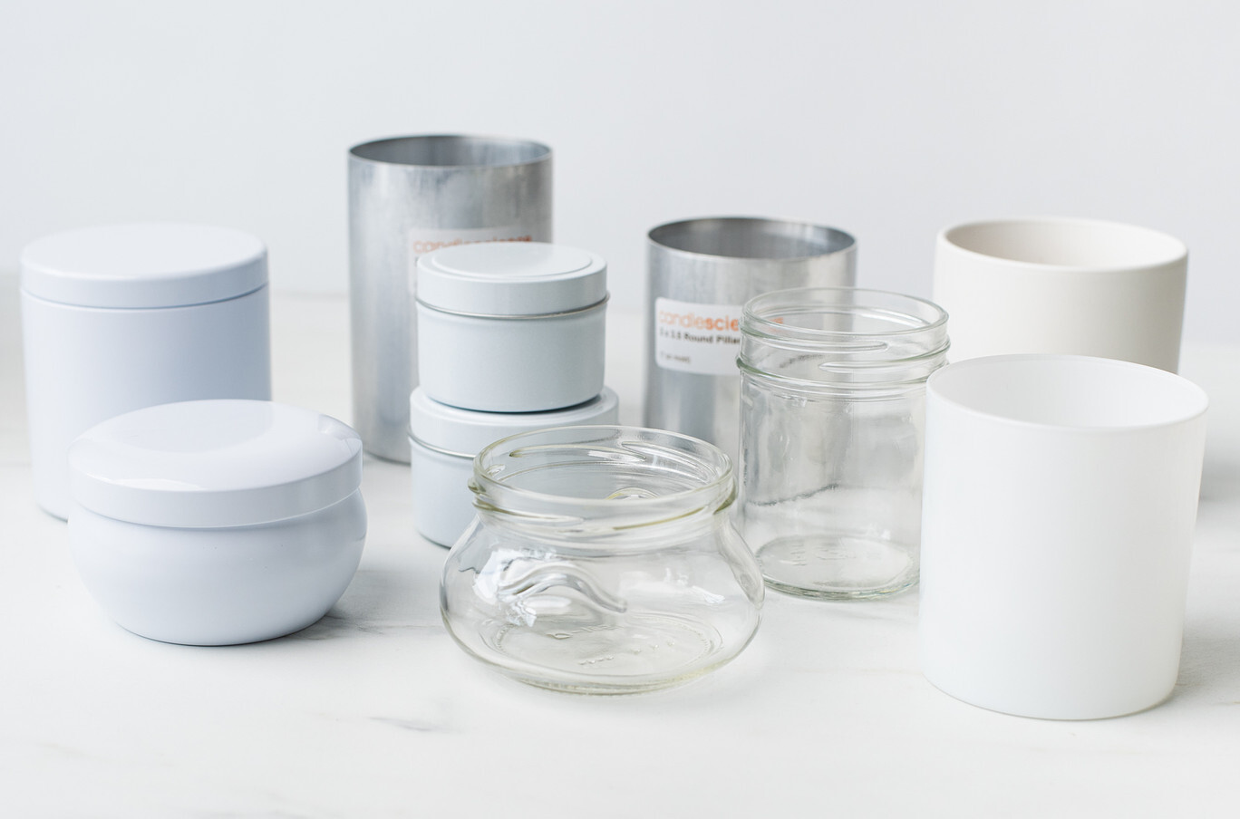 An array of candle containers, including a white glass jar, clear glass jars, a white ceramic jar, white tins, and silver pillar candle molds, sit against a white background.