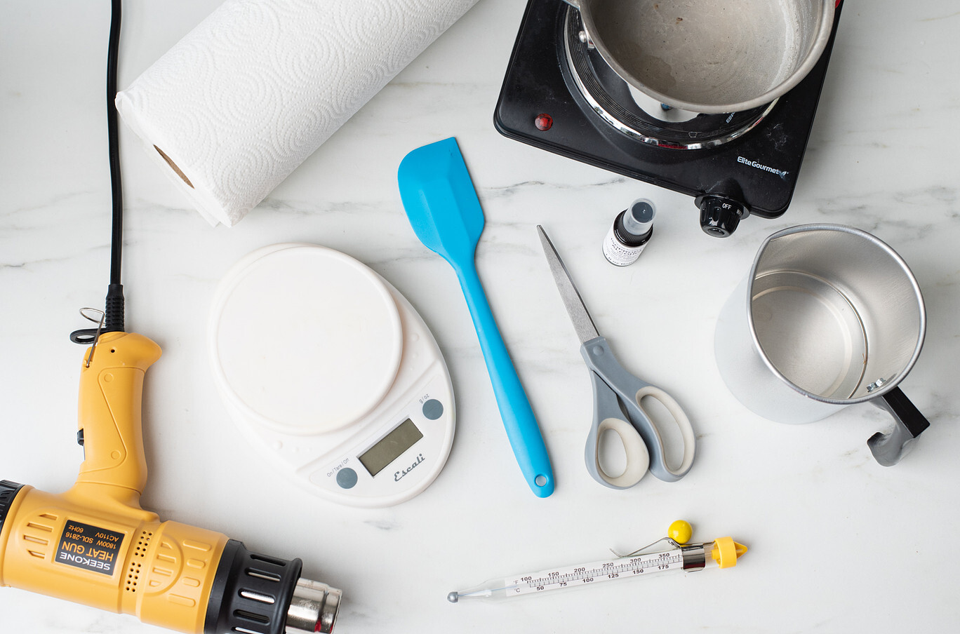 An overhead view of candle making equipment, including a yellow heat gun, glass candy thermometer, digital scale, silicone spatula, scissors, a roll of paper towels, a spritz bottle of isopropyl alcohol, a metal pouring pitchers, and a hot plate.