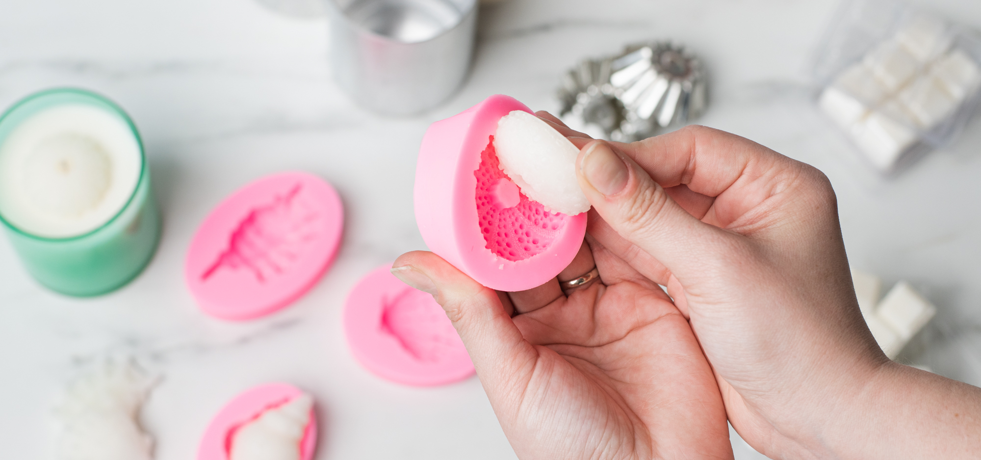 A marble surface with pink seashell-shaped wax melt molds, silver cupcake-shaped wax melt molds, and white wax melts scattered throughout. 