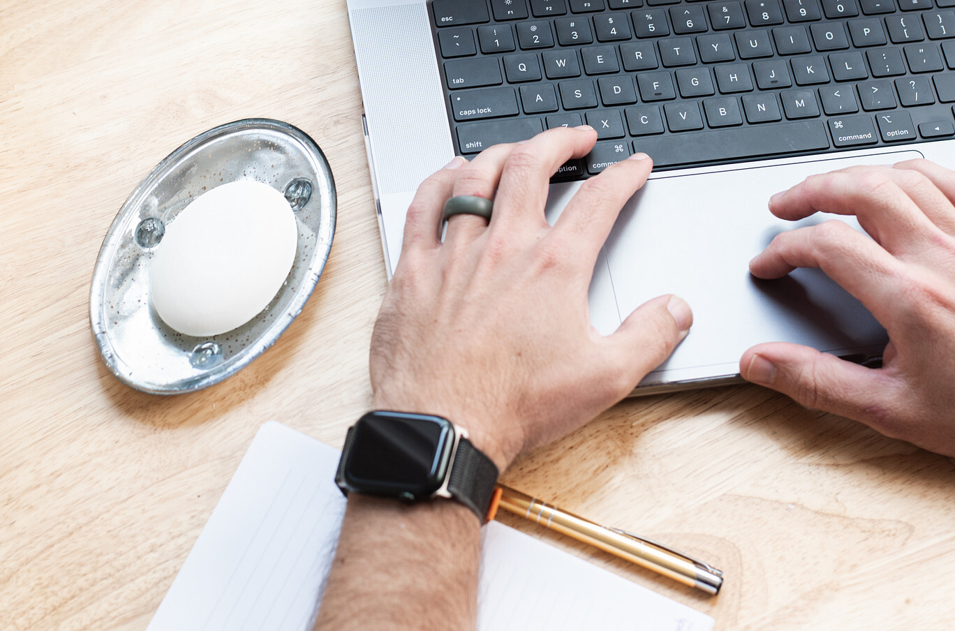 A pair of light colored hands types on a laptop computer; a bar of white soap in a silver dish sits next to the laptop