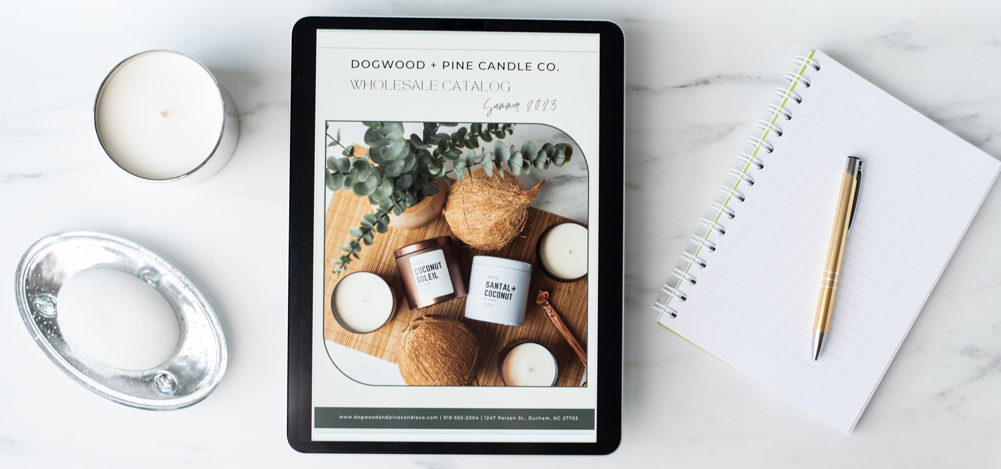 A flat lay of an iPad displaying a wholesale catalog for Dogwood + Pine Candle Co. Flanking the iPad to the left is a white bar of soap in a silver dish and a white soy candle in a silver tumbler jar. To the right of the iPad is a lined notebook and gold pen.