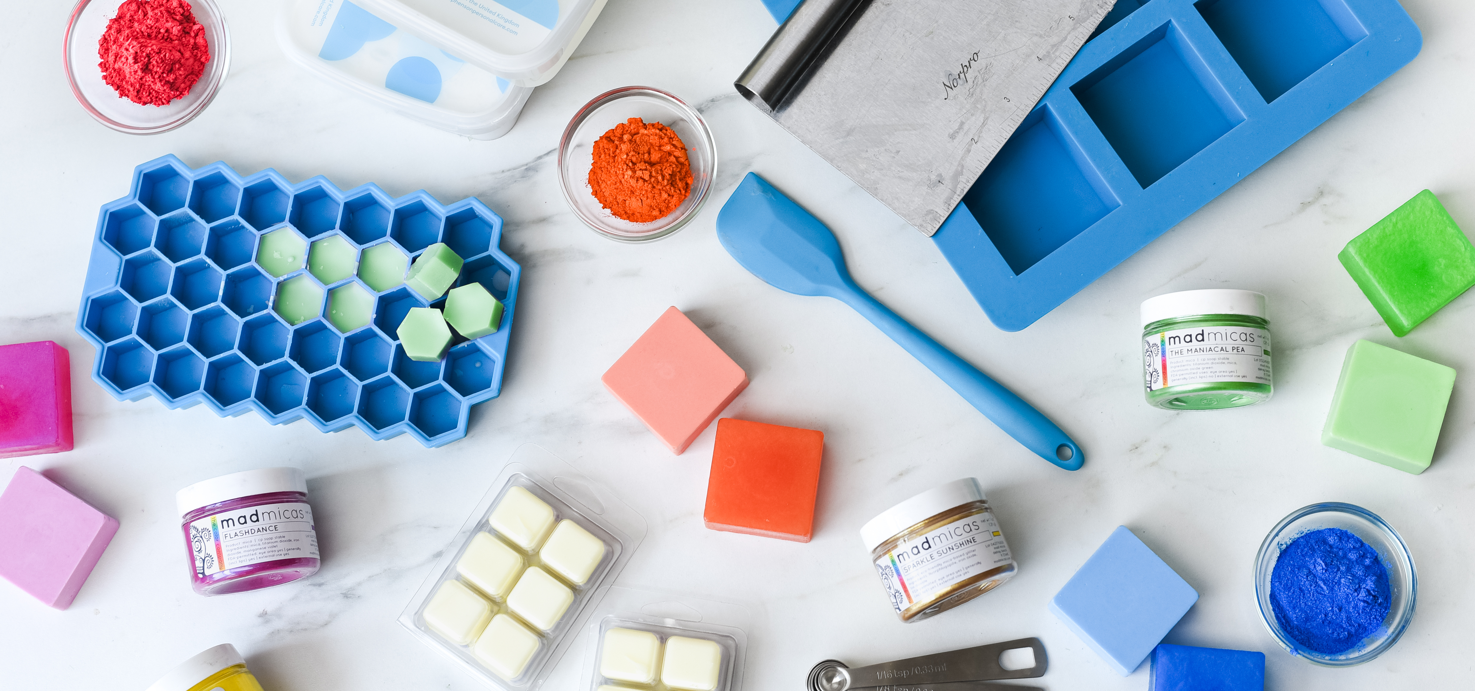 A white marble surface with glass bowls filled with pink, orange, and blue mica powders, jars of fuchsia, green, and gold mica powders with labels that say "mad micas," and soap and wax melts that match the mica powders.