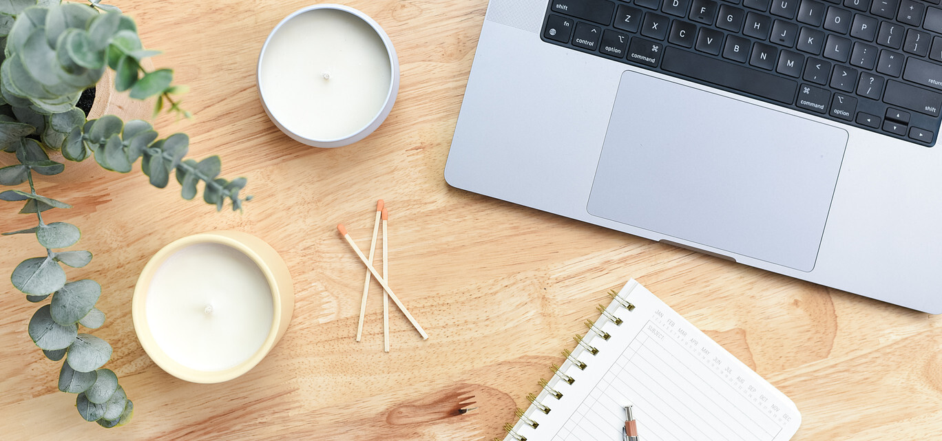 An overhead view of a natural wood desk; on the desk is a succulent plant and two candles, plus three matchsticks, a silver laptop computer, and a paper planner with a pen