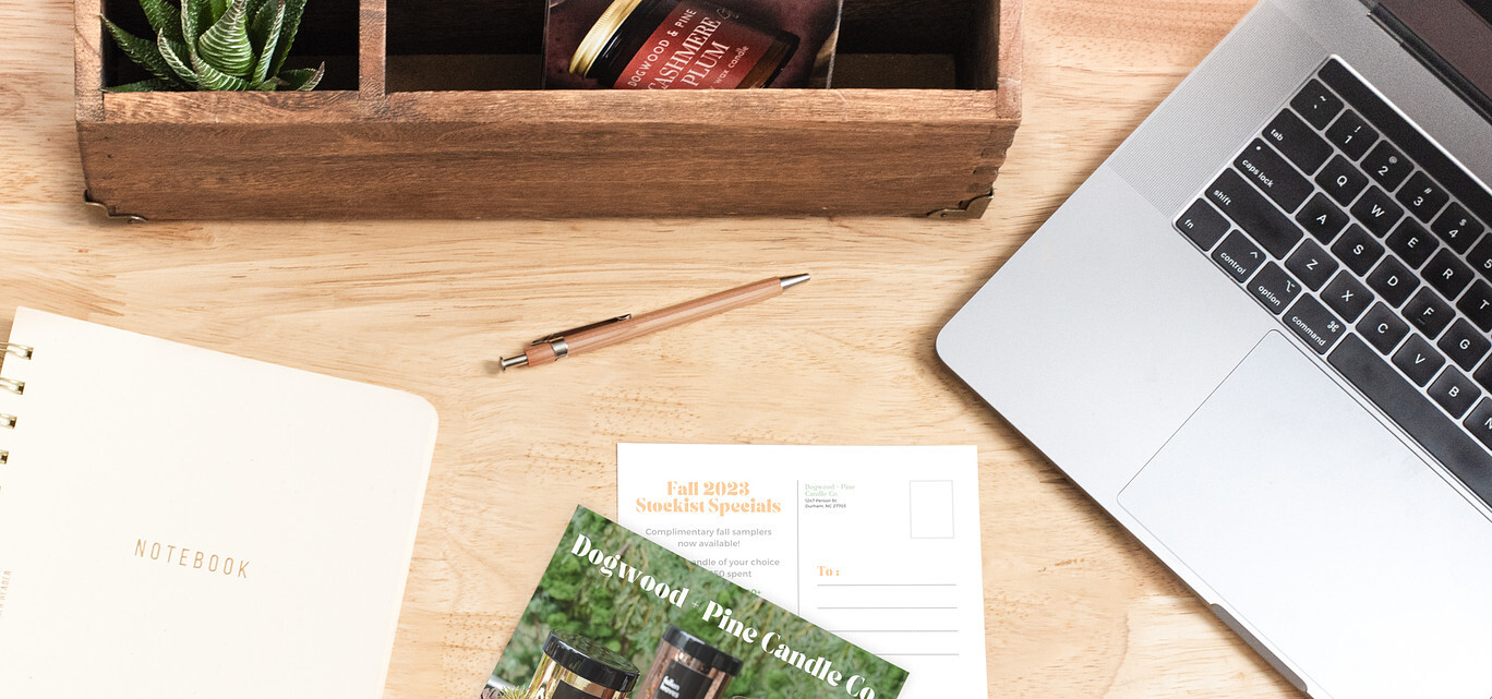 An overhead view of a natural wood desk; on the desk is a a silver laptop computer, notebook, natural wood pen, wholesale postcards, and a wooden box holding a succulent plant and more wholesale pitch postcards