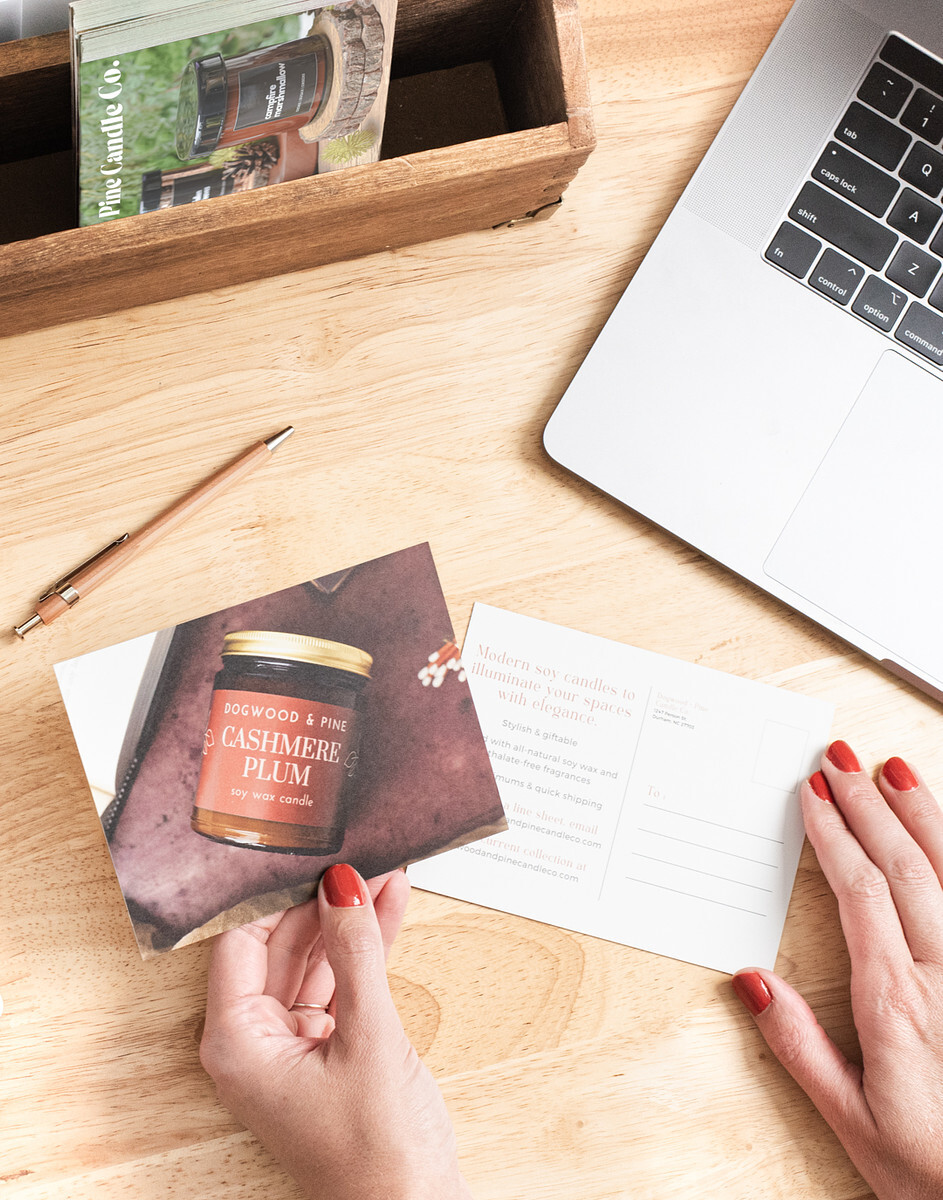 An overhead view of a woman's hand with painted nails holding a wholesale pitch postcard with an image of candle in an amber jar with gold lid, and a label that reads Cashmere Plum. Behind the postcard is a natural wood-colored desktop with a silver laptop, and a wooden desk organizer holding more wholesale postcards.