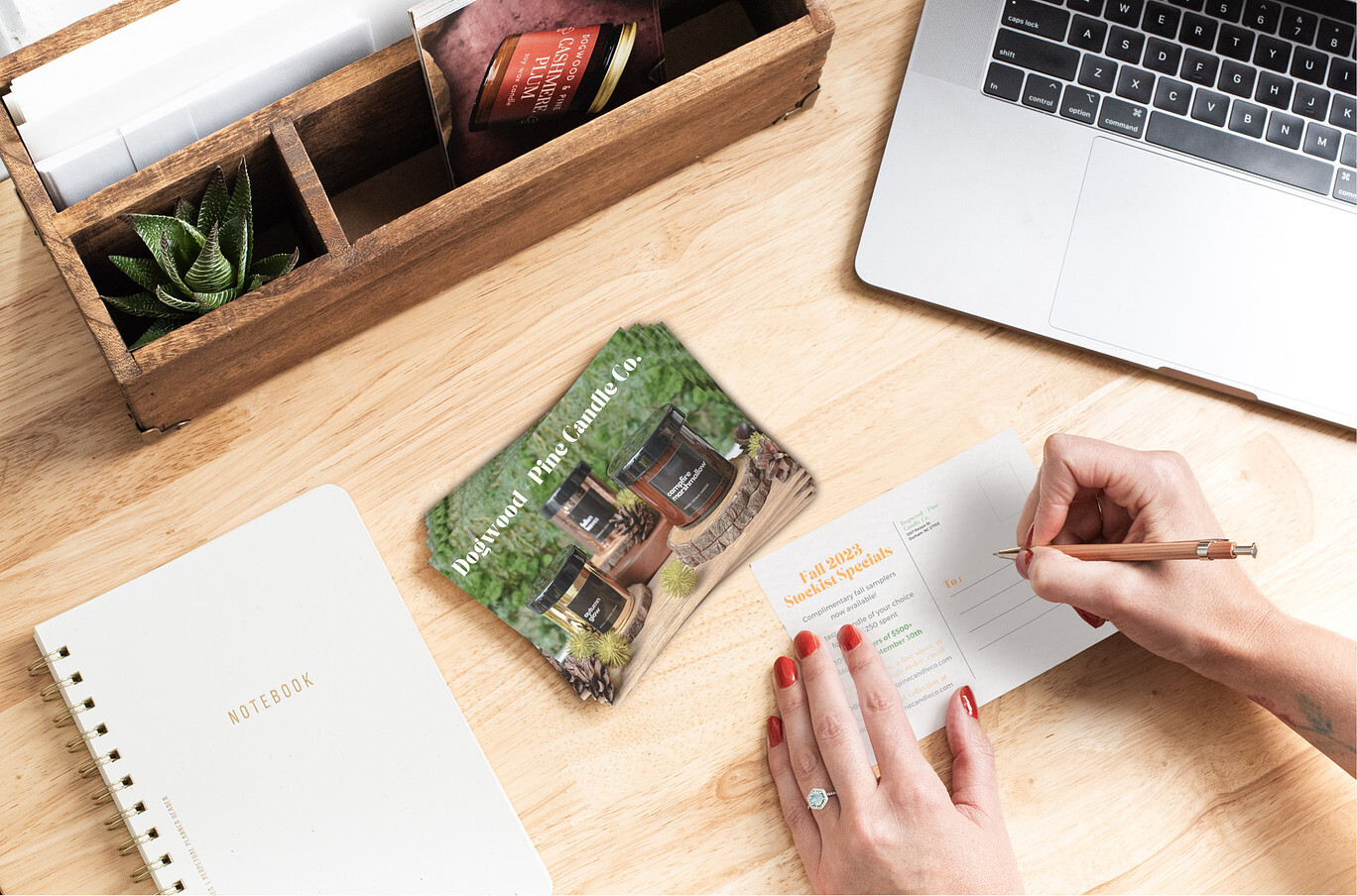 An overhead view of a natural-colored wood desk. To the left is a white, spiral-bound notebook; in the center is a pair of women's hands addressing a wholesale pitch postcard. Rounding out the scent is a silver laptop and a wooden desk organizer with more wholesale postcards tucked in it.