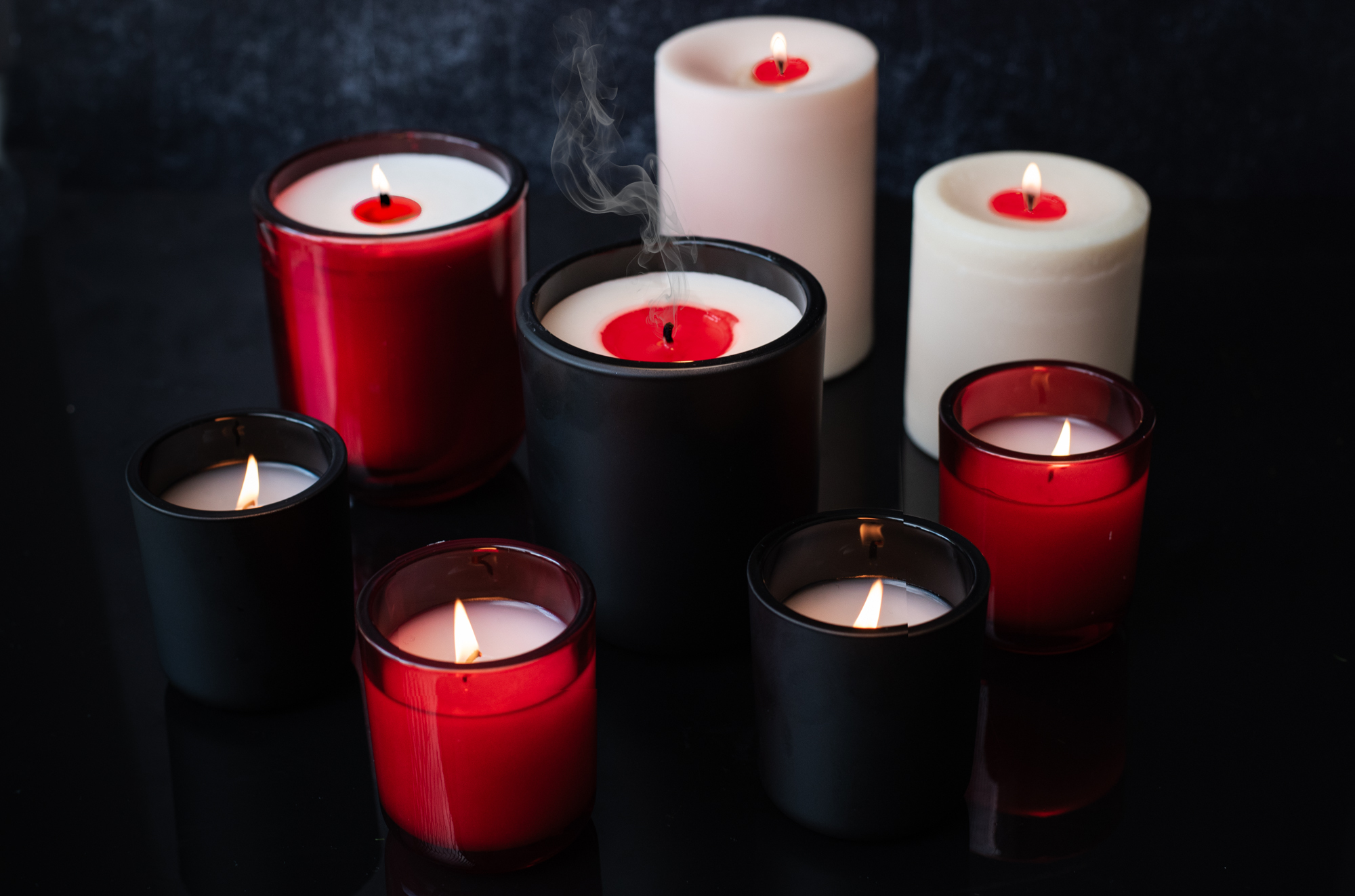 A group of lit candles made in matte black jars, red glass jars, and pillars, some revealing red wax as they burn.