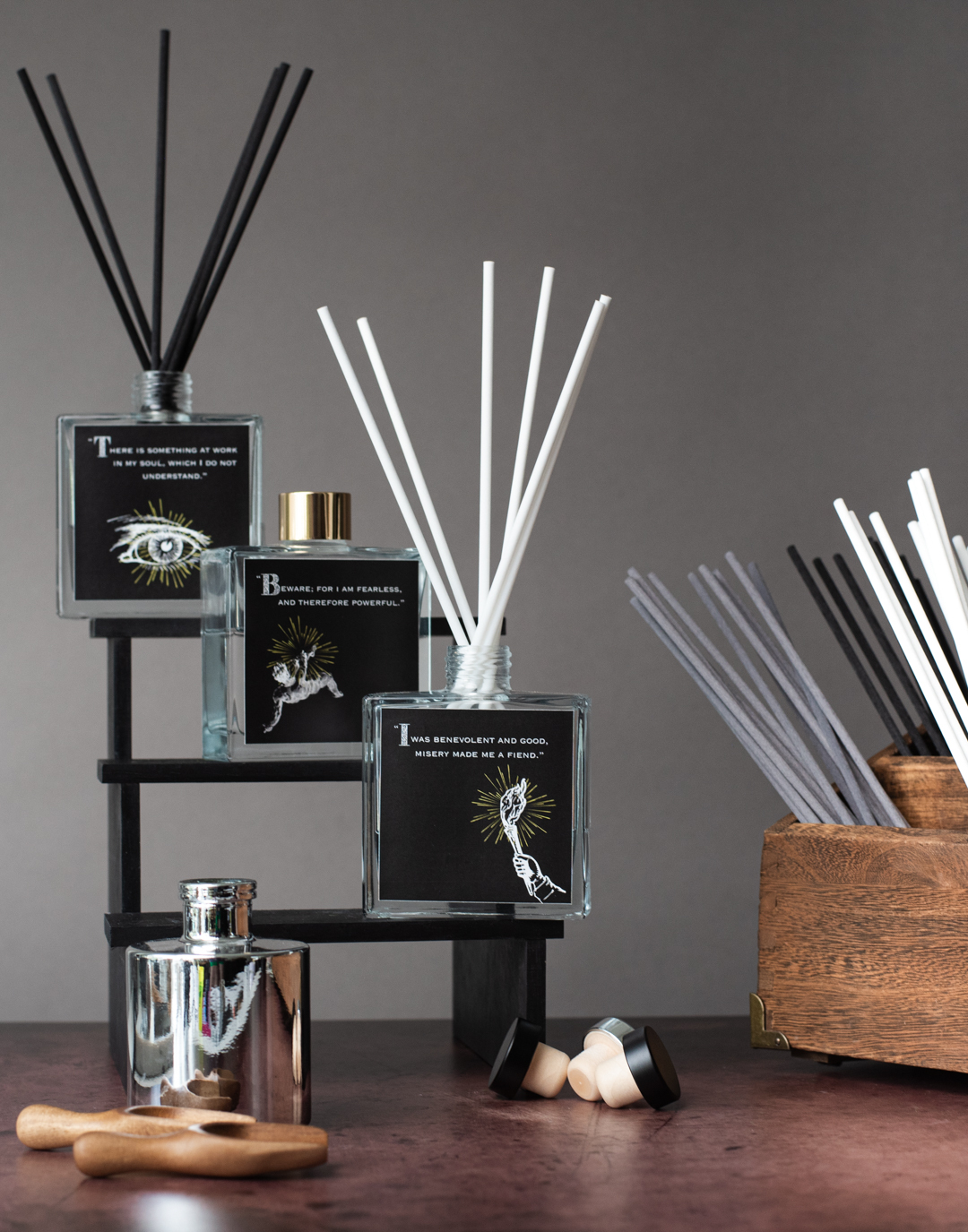An image of three square glass reed diffusers with black and white reeds. Their labels are black and white and feature quotes from Mary Shelley's novel, Frankenstein. More black, white, and gray reeds are features off to the side out of frame in a wooden box.