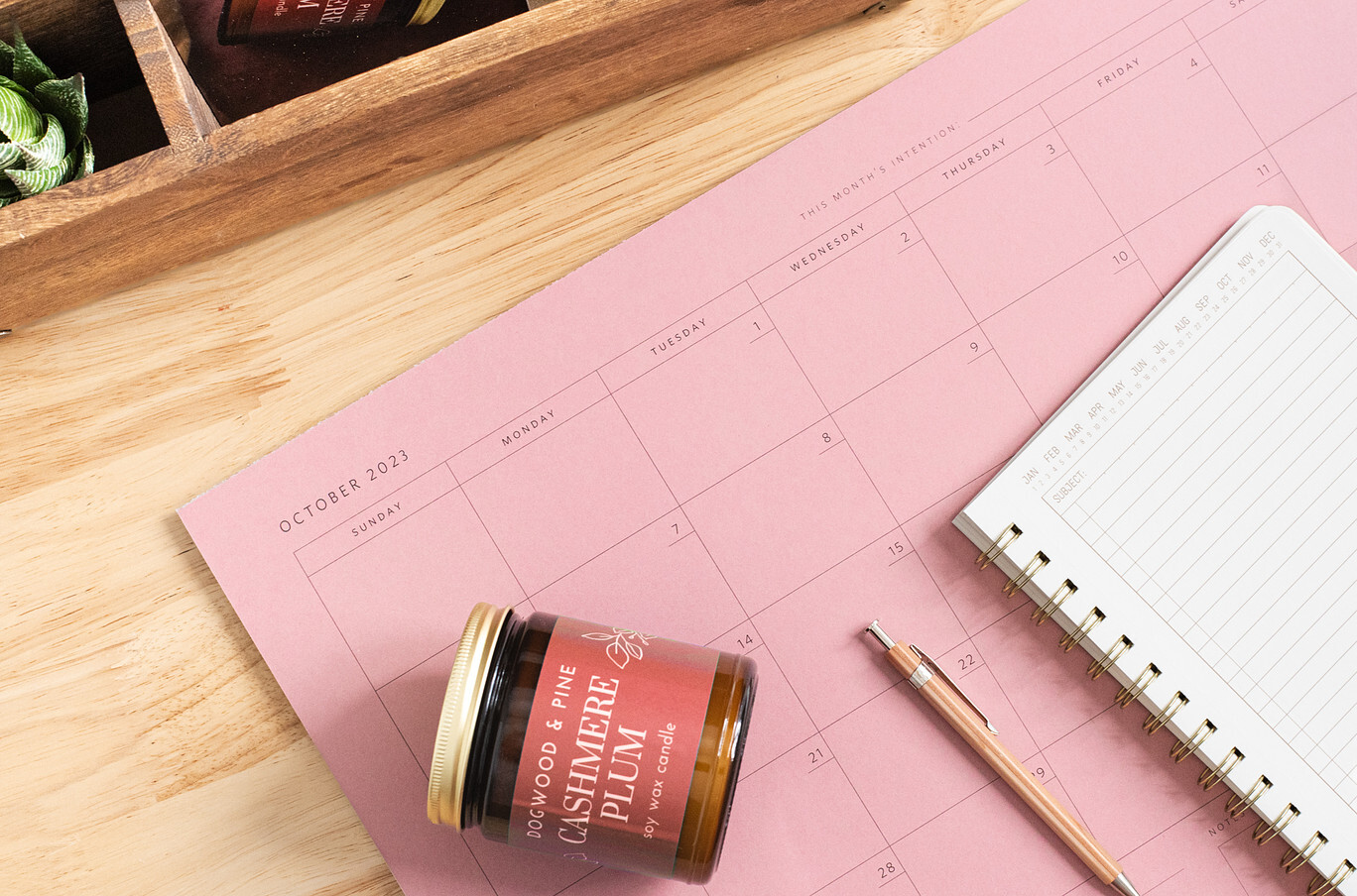 An overhead view of a natural-colored wood desktop. On the desk is a Cashmere Plum candle in a glass amber jar with gold lid; a month-view calendar, a notebook planner, and a wooden pencil.