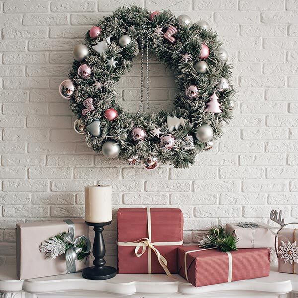 White brick fireplace and mantel are decorated for the holidays with a hanging wreath on the brick, presents wrapped in red paper. A white pillar candle sits atop a dark stand on the mantel.