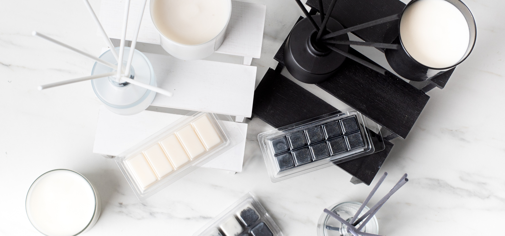 An assortment of black and white reed diffusers, wickless candles, and wax melts on a white marble surface.